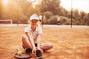 si siede su il terra. giovane femmina tennis giocatore nel allegro Abiti è su il Tribunale all'aperto foto