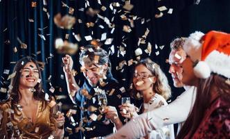 coriandoli è nel il aria. gruppo di allegro amici festeggiare nuovo anno in casa con bevande nel mani foto