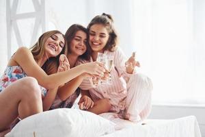 festeggiare con bicchieri di alcool nel mani. contento femmina amici avendo bene tempo a pigiama festa nel il Camera da letto foto