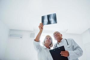 anziano uomo e donna medici nel bianca uniforme esamina raggi X di umano gambe foto