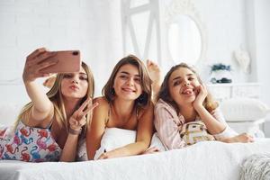 fabbricazione autoscatto di utilizzando rosa Telefono. contento femmina amici avendo bene tempo a pigiama festa nel il Camera da letto foto