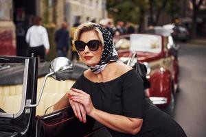 bionda donna nel occhiali da sole e nel nero vestito pendente su vecchio Vintage ▾ classico auto foto