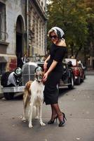 bionda donna nel occhiali da sole e nel nero vestito vicino vecchio Vintage ▾ classico auto con sua cane foto