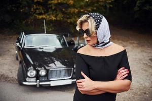 bionda donna nel occhiali da sole e nel nero vestito vicino vecchio Vintage ▾ classico auto foto