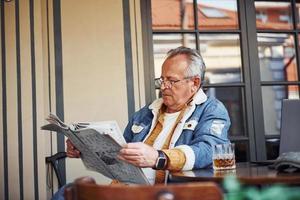 elegante anziano nel alla moda Abiti e nel bicchieri si siede nel il bar e legge giornale foto