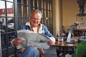 elegante anziano nel alla moda Abiti e nel bicchieri si siede nel il bar e legge giornale foto