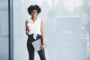 giovane africano americano donna nel bicchieri con bloc notes e tazza di bevanda nel mani sta in casa foto