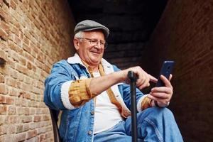 elegante anziano nel alla moda Abiti e nel bicchieri si siede su il sedia e usi Telefono foto
