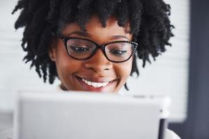 giovane africano americano donna nel bicchieri sta in casa nel il ufficio con tavoletta nel mani foto