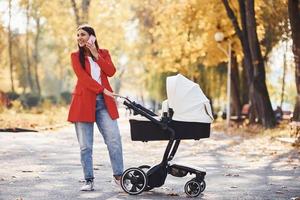 parlando di il Telefono. madre nel rosso cappotto avere un' camminare con sua ragazzo nel il carrozzina nel il parco a autunno tempo foto