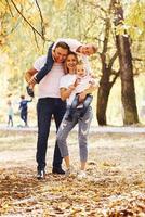 madre e papà detiene bambini a le spalle e nel mani. allegro giovane famiglia avere un' camminare nel un autunno parco insieme foto
