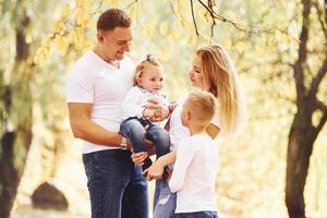 madre e papà detiene bambini a le spalle e nel mani. allegro giovane famiglia avere un' camminare nel un autunno parco insieme foto