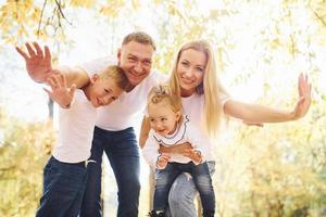 madre e papà detiene bambini a le spalle e nel mani. allegro giovane famiglia avere un' camminare nel un autunno parco insieme foto