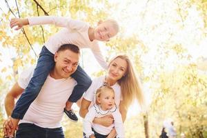 madre e papà detiene bambini a le spalle e nel mani. allegro giovane famiglia avere un' camminare nel un autunno parco insieme foto