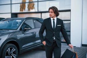 ritratto di bello giovane uomo d'affari nel nero completo da uomo e cravatta all'aperto vicino moderno auto e con shopping borse foto