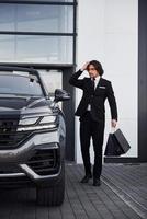 ritratto di bello giovane uomo d'affari nel nero completo da uomo e cravatta all'aperto vicino moderno auto e con shopping borse foto