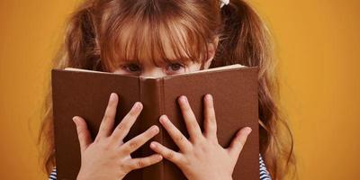 carino poco ragazza con libro nel mani nel il studio contro giallo sfondo foto