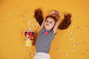 carino poco ragazza con Popcorn dire bugie giù su il giallo pavimento foto