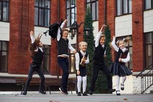 gruppo di bambini nel scuola uniforme salto e avendo divertimento all'aperto insieme vicino formazione scolastica edificio foto