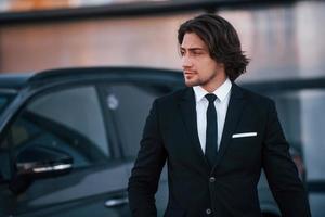ritratto di bello giovane uomo d'affari nel nero completo da uomo e cravatta all'aperto vicino moderno auto foto