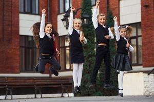 gruppo di bambini nel scuola uniforme salto e avendo divertimento all'aperto insieme vicino formazione scolastica edificio foto