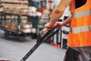 giovane maschio lavoratore nel uniforme è nel il magazzino spingendo pallet camion foto