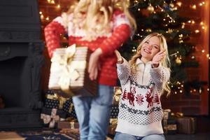 poco ragazza in piedi con regalo scatola e fa sorpresa per madre in casa a Natale vacanze tempo foto