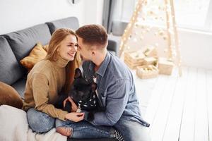 contento giovane moderno coppia seduta su il divano a casa con Natale albero con loro carino cane foto