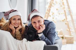 contento giovane moderno coppia nel Natale cappelli dire bugie giù a divano a casa con loro carino cane foto