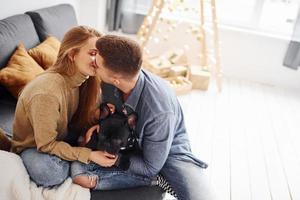 contento giovane moderno coppia seduta su il divano a casa con Natale albero con loro carino cane foto