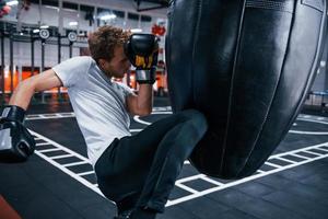 giovane uomo nel bianca camicia e boxe protettivo guanti fare esercizi nel Palestra con spingendo Borsa foto