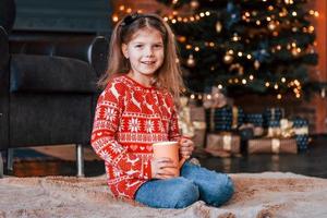 carino poco ragazza seduta su il terra con tazza di bevanda nel il Natale decorato camera foto