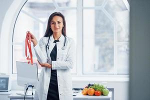 femmina dietologo nel bianca cappotto in piedi in casa nel il ufficio a posto di lavoro con misurazione nastro nel mani foto