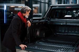 alla moda vecchio uomo con grigio capelli e baffi guardare per un' nuovo auto in casa nel salone foto