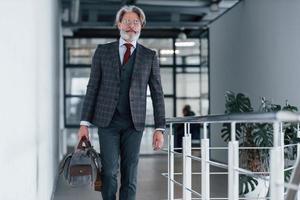 anziano uomo d'affari nel completo da uomo e cravatta con grigio capelli e barba a piedi in casa con Borsa foto