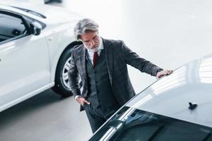 aereo Visualizza di alla moda vecchio uomo con grigio capelli e baffi quello guardare per un' nuovo auto in casa nel salone foto