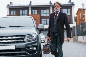 alla moda anziano uomo con grigio capelli e barba passeggiate con Borsa all'aperto su il strada vicino il suo auto foto