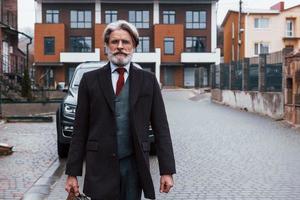 alla moda anziano uomo con grigio capelli e barba passeggiate con Borsa all'aperto su il strada vicino il suo auto foto