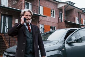 alla moda anziano uomo con grigio capelli e barba avere conversazione di Telefono all'aperto su il strada vicino il suo auto foto