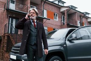 alla moda anziano uomo con grigio capelli e barba avere conversazione di Telefono all'aperto su il strada vicino il suo auto foto