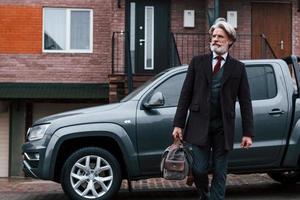 alla moda anziano uomo con grigio capelli e barba passeggiate con Borsa all'aperto su il strada vicino il suo auto foto