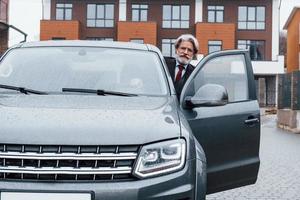 alla moda anziano uomo con grigio capelli e barba seduta nel il suo moderno nuovo auto su il strada foto