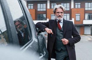 alla moda anziano uomo con grigio capelli e barba pendente su il suo auto e Tenere chiavi foto