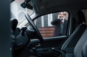 alla moda anziano uomo con grigio capelli e barba avere conversazione di Telefono all'aperto su il strada vicino il suo auto foto