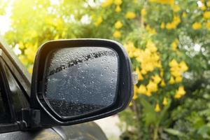 vicino su su il copertina plastica di specchio ala di auto con su il bicchiere con far cadere di acqua pioggia. morbido leggero nel il giardino con luminosa giallo fiore di giallo Sambuco. foto