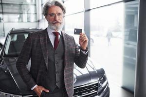 elegante anziano uomo con grigio capelli e barba sta contro moderno nero auto con credito carta nel mano foto