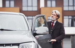 alla moda anziano uomo con grigio capelli e barba è all'aperto su il strada vicino il suo auto apertura il porta foto