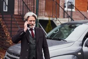alla moda anziano uomo con grigio capelli e barba è all'aperto su il strada vicino il suo auto avere conversazione di il Telefono foto