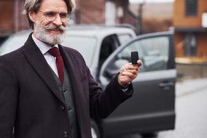 alla moda anziano uomo con grigio capelli e barba in piedi all'aperto su il strada vicino il suo auto con chiavi nel mano foto