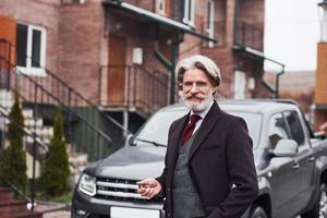 alla moda anziano uomo con grigio capelli e barba è all'aperto su il strada vicino il suo auto foto